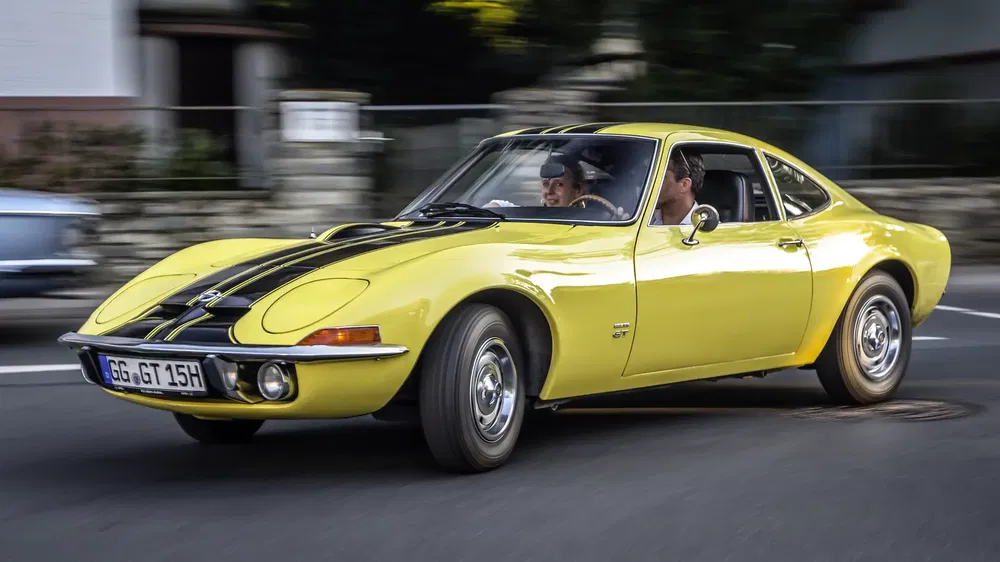 Klassieke gele Opel GT met zwarte strepen, rijdend op een weg met passagiers erin.