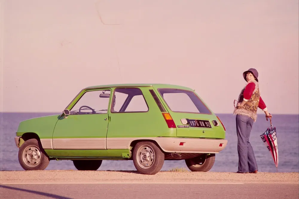 Voiture Renault 5 verte stationnée près de la mer avec une personne tenant un parapluie.