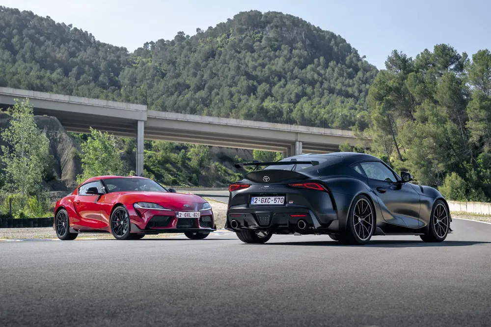 Twee Toyota Supra's, rood en zwart, geparkeerd voor een schilderachtig berglandschap.