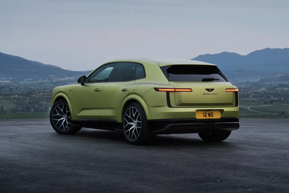 SUV Bentley Bentayga vert sur route panoramique, montagnes en arrière-plan sous ciel nuageux.