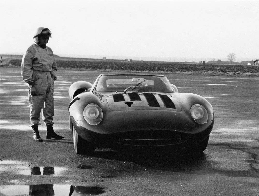Homme en combinaison de course avec une Jaguar XJ13 sur une piste humide.