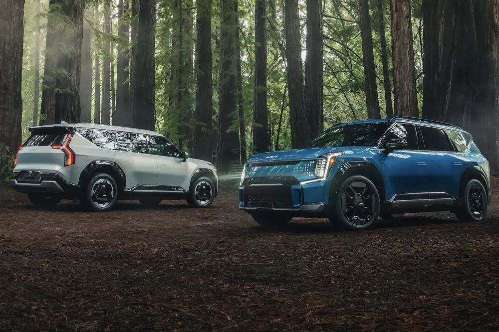 Twee Kia EV9 elektrische auto's geparkeerd in een bos, modellen in blauw en wit.