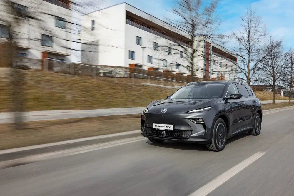 MG ZS EV roulant sur une route urbaine, bâtiments modernes en arrière-plan.