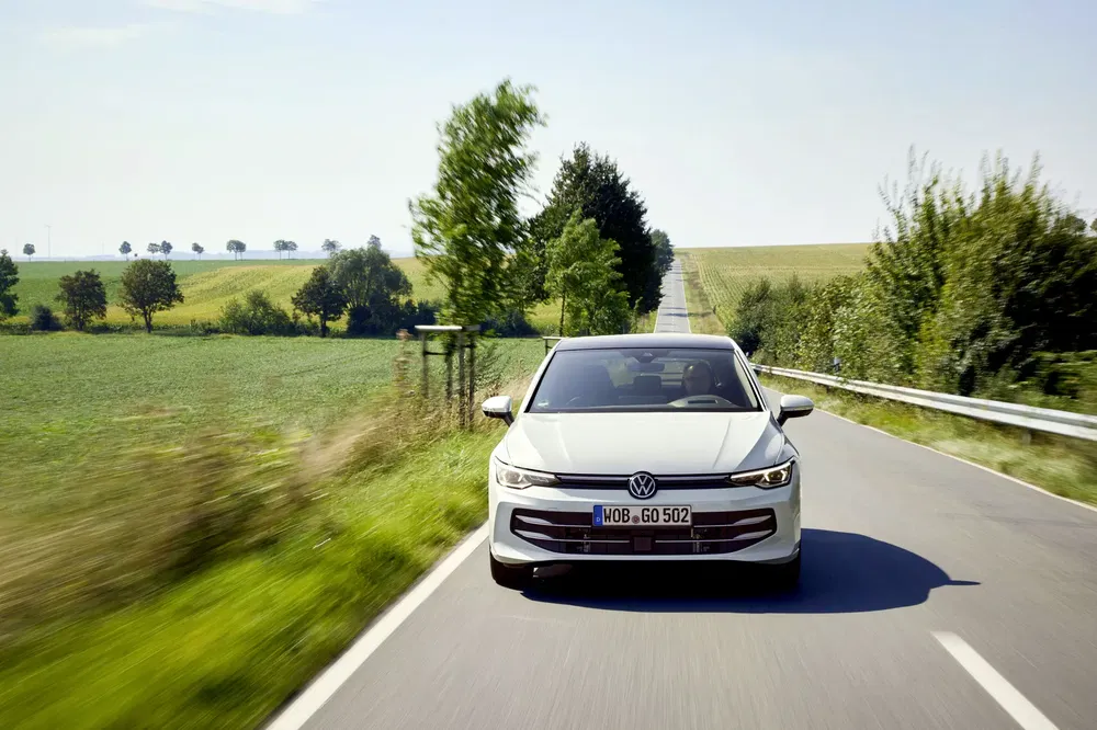 Witte Volkswagen Golf rijdt op landelijke weg omgeven door groene velden.