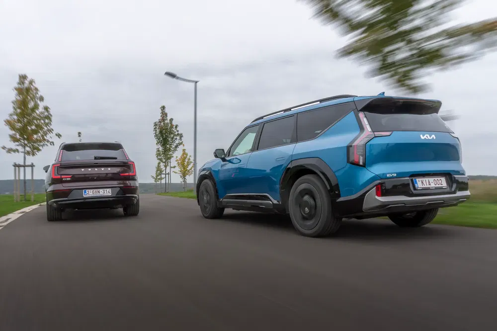 Kia EV9 bleu et SUV noir roulant sur une route avec arbres en arrière-plan.