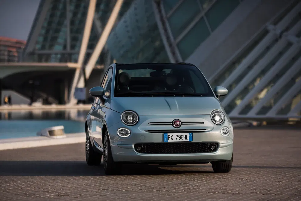 Lichtblauwe Fiat 500 voor modern gebouw, avondlicht schijnt op de auto.
