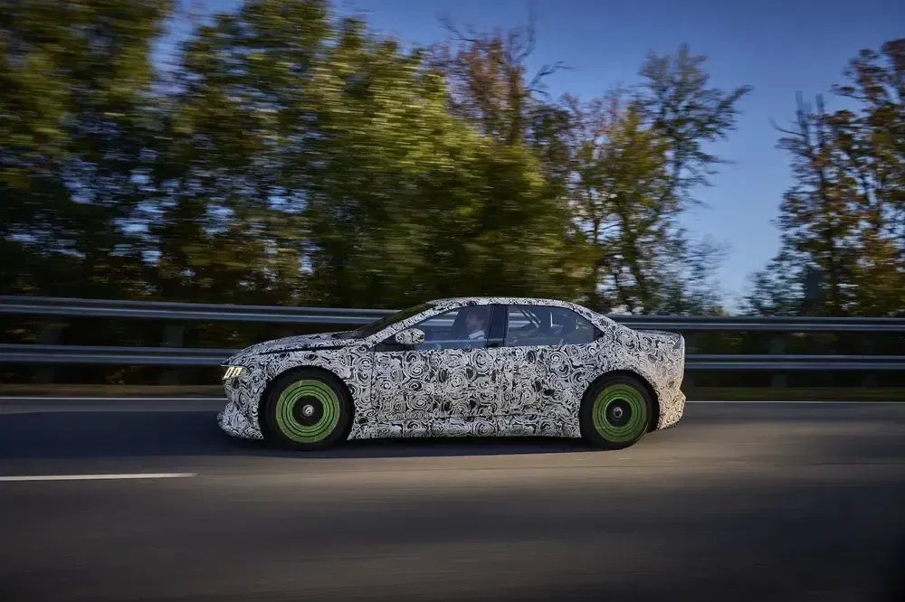 BMW i5 prototype met camouflage rijdt langs een met bomen omzoomde weg.