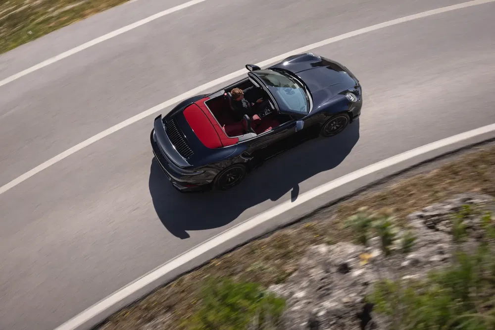 Porsche cabriolet noir sur route sinueuse vue aérienne, capote rouge, vitesse et élégance.