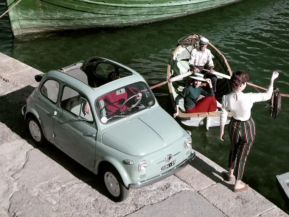 Groene vintage Fiat 500 geparkeerd bij een kade met een kleine roeiboot.