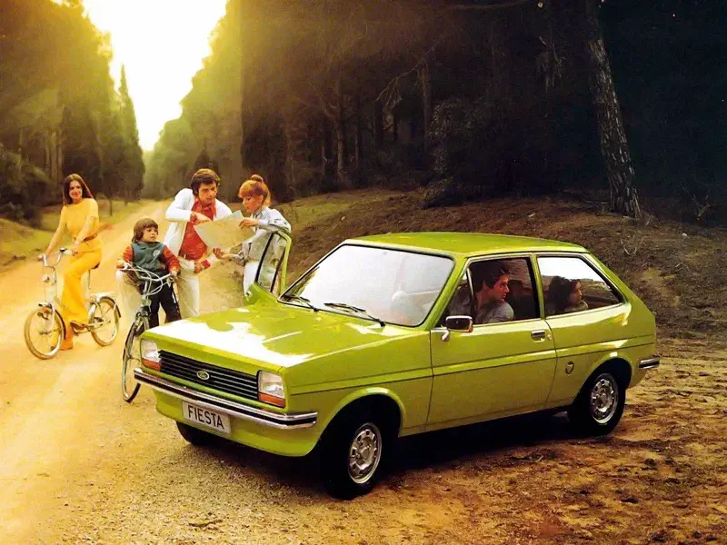 Ford Fiesta verte des années 70 sur un chemin forestier avec une famille à proximité.