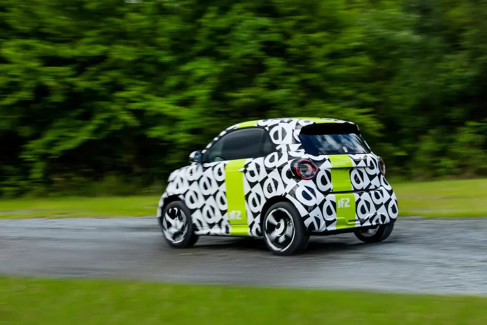 Voiture compacte Smart #2 camouflée, roulant sur une route bordée d'arbres verts.