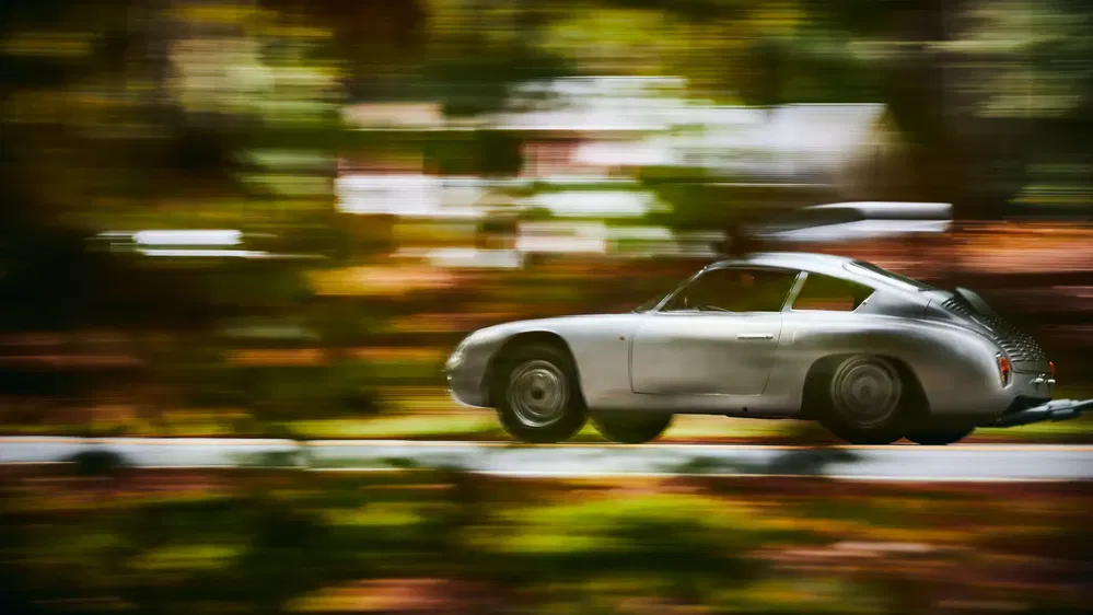 Voiture classique argentée roulant à grande vitesse sur une route bordée d'arbres flous.