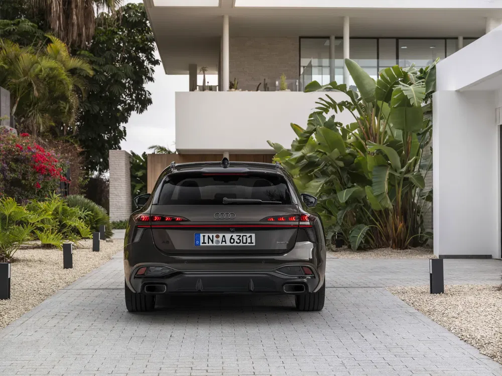 Audi A6 noir garée devant une maison moderne avec végétation tropicale.