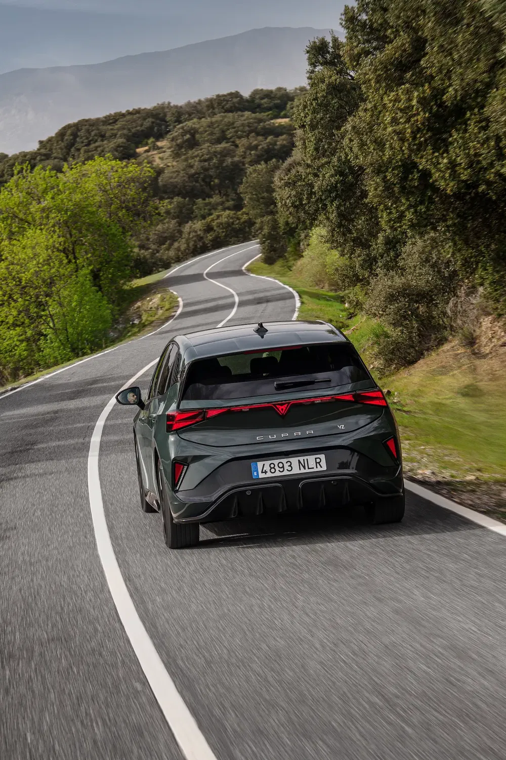 Cupra compact moderne sur route sinueuse entourée de forêts et collines verdoyantes.
