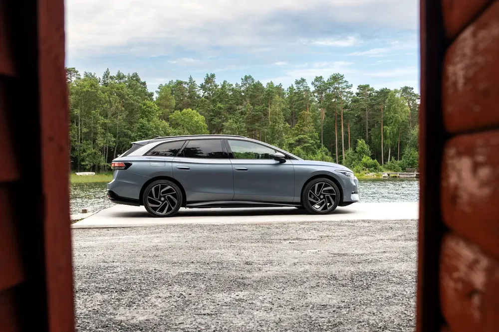 Break électrique Volkswagen ID.7 gris foncé près d'un lac entouré de forêts verdoyantes.
