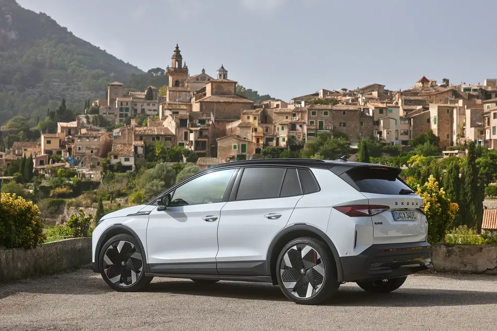 Skoda Enyaq iV blanc garé devant un village pittoresque, paysage montagneux à l'arrière-plan.