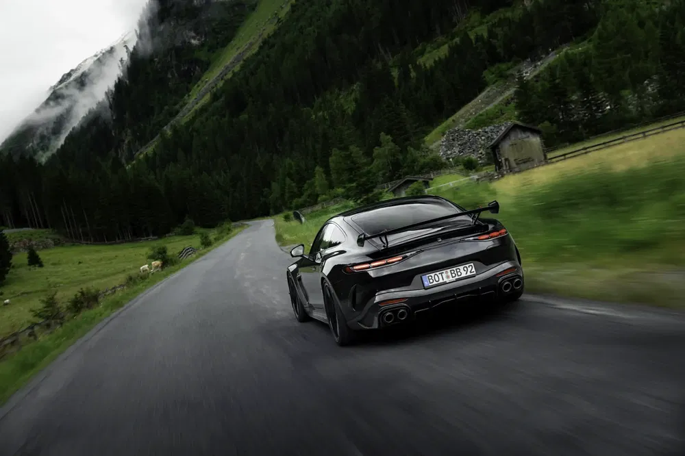 Porsche 911 Turbo noir roulant sur une route de montagne sinueuse, décor alpin verdoyant.