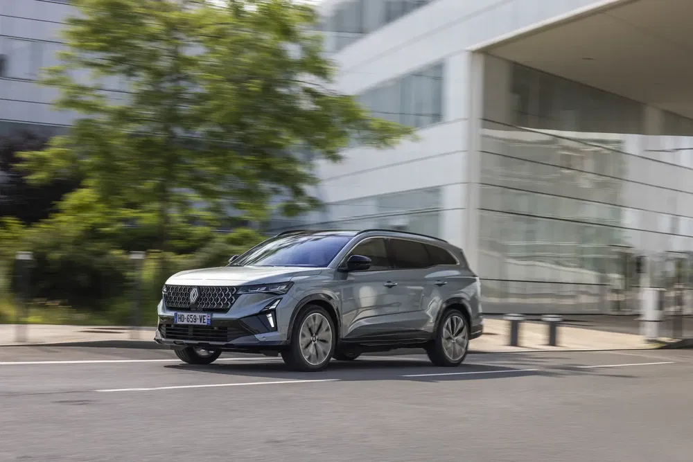 Grijze Renault Espace SUV rijdt voor een modern gebouw langs.