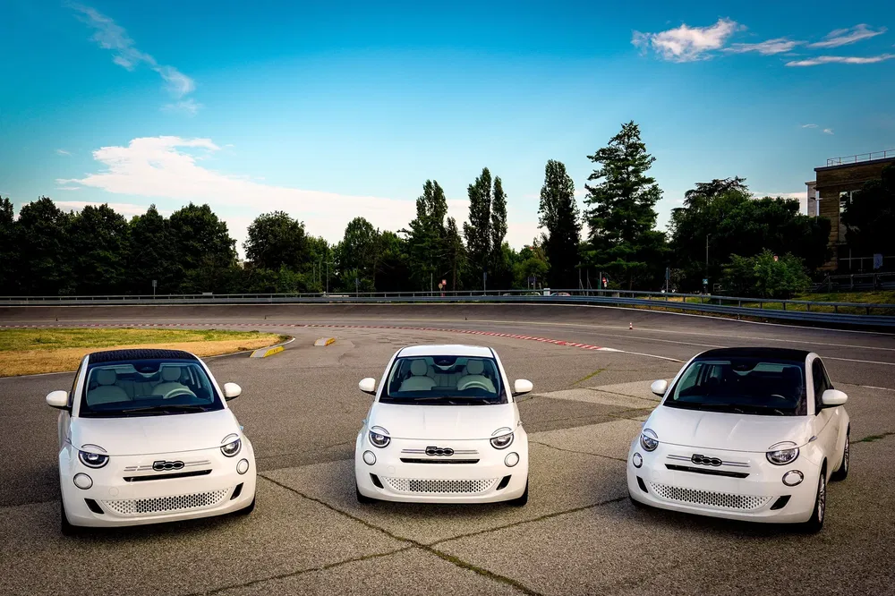 Drie witte Fiat 500's naast elkaar op een parkeerplaats, omringd door bomen.