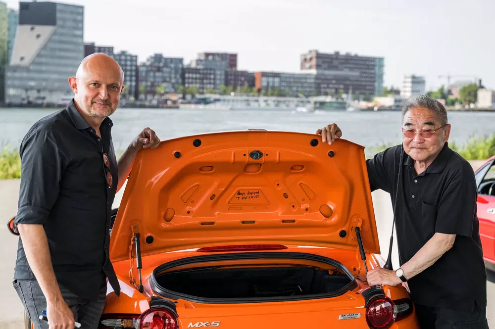 Oranje Mazda MX-5 met open kofferbak, twee mannen poseren ernaast, stedelijke achtergrond.