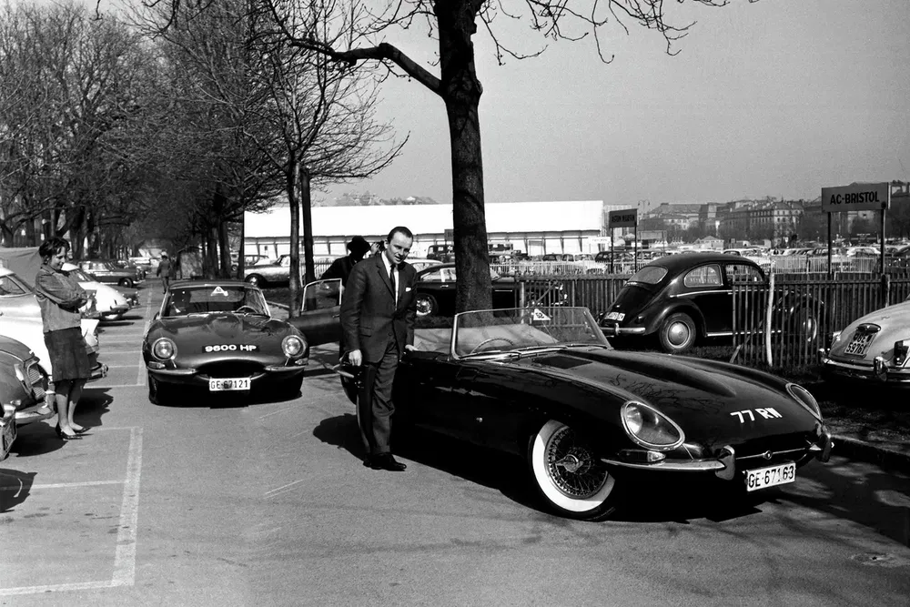 Zwarte Jaguar E-Type geparkeerd, man in pak ernaast, klassieke auto's op achtergrond.