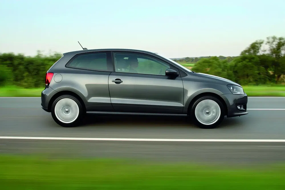 Grijze Volkswagen Polo rijdt op weg met groene achtergrond.