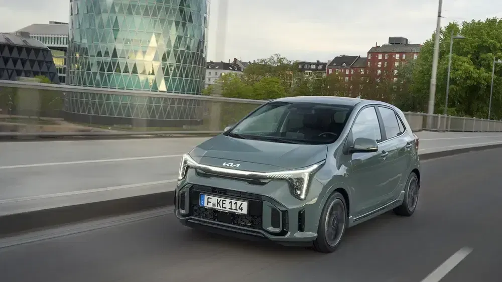 Kia Picanto verte traverse un pont urbain, devant un bâtiment moderne en verre.