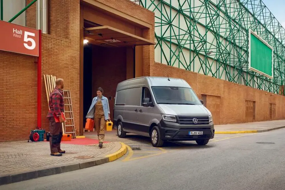 Fourgon Volkswagen gris garé devant un entrepôt, avec ouvriers échangeant à l'entrée.