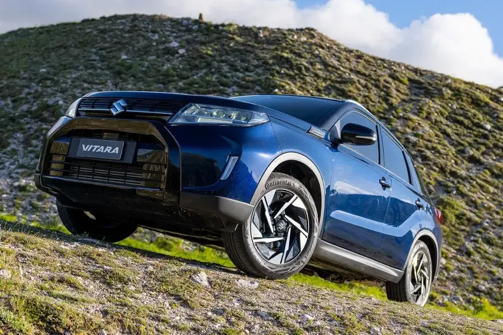 Suzuki Vitara bleu sur terrain vallonné, pneus Continental visibles, ciel partiellement nuageux.