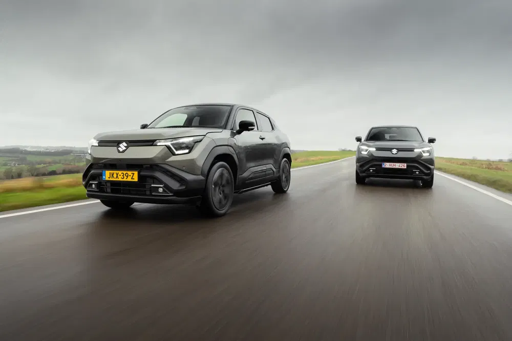 Twee grijze Suzuki SUV's rijden op een natte weg onder een bewolkte hemel.
