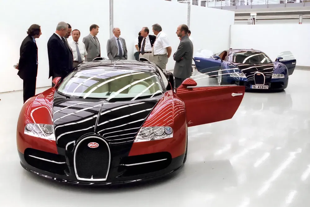{"fr":"Bugatti Veyron rouge et noire exposée, entourée de spectateurs intéressés dans un showroom moderne.","nl":"Rode en zwarte Bugatti Veyron in showroom met belangstellende kijkers."}