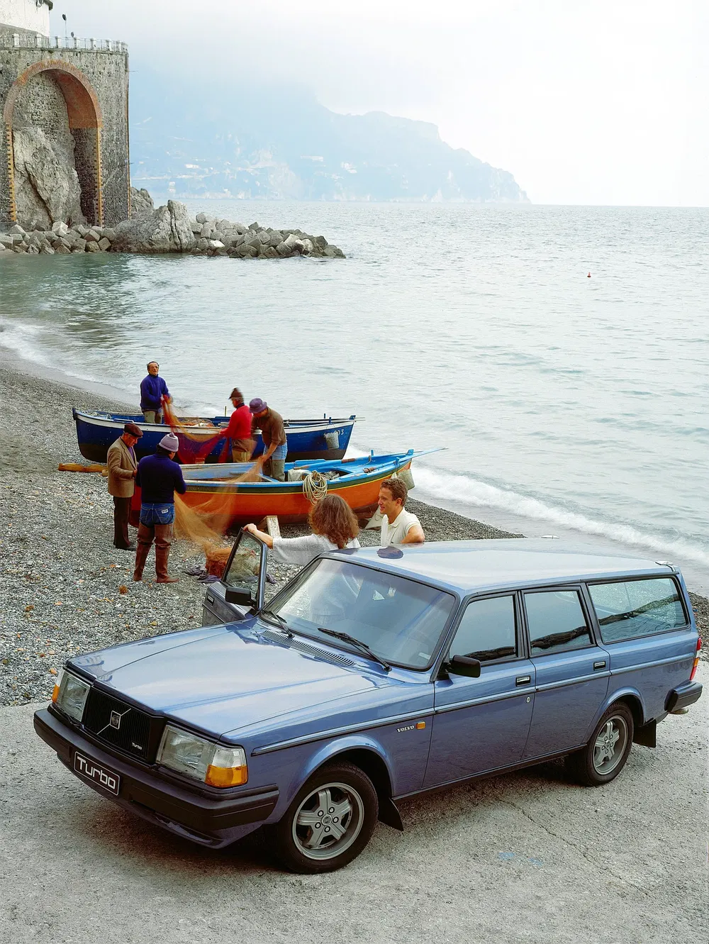 Une Volvo 240 Turbo bleue garée sur une plage rocheuse avec des pêcheurs à l'arrière-plan.