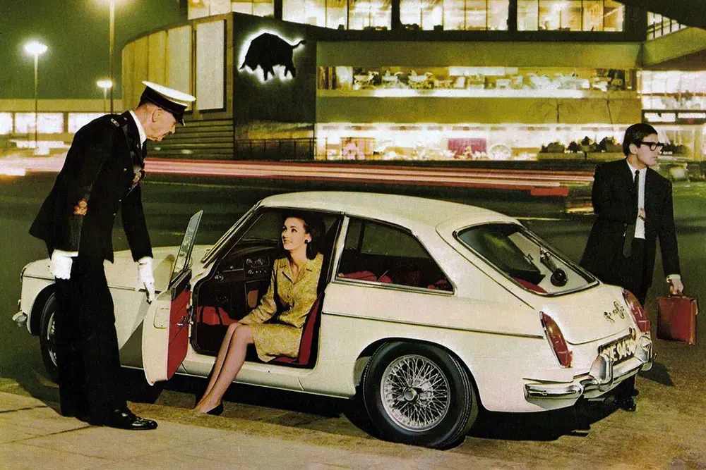 Voiture sportive classique stationnée devant un bâtiment moderne, passager assisté par un portier.