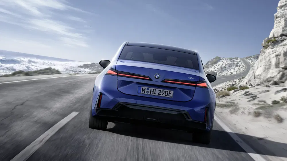 Achterkant van een blauwe BMW iX3 op een zonnige, bochtige kustweg.