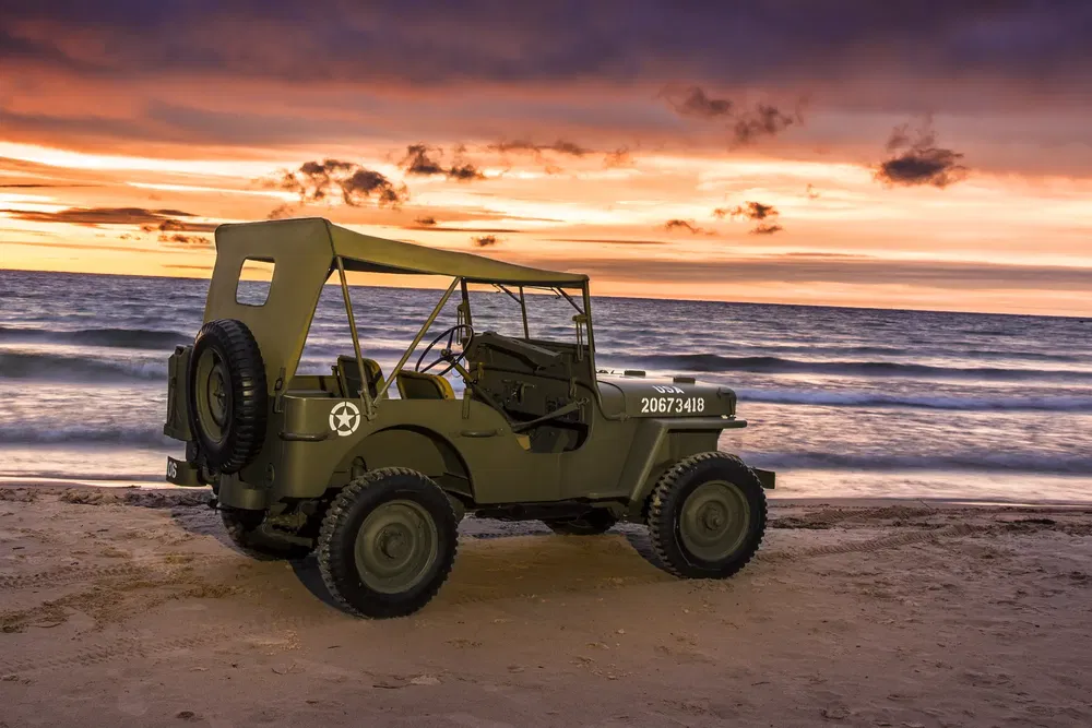 Vintage militaire Jeep op strand bij zonsondergang met zee op de achtergrond.