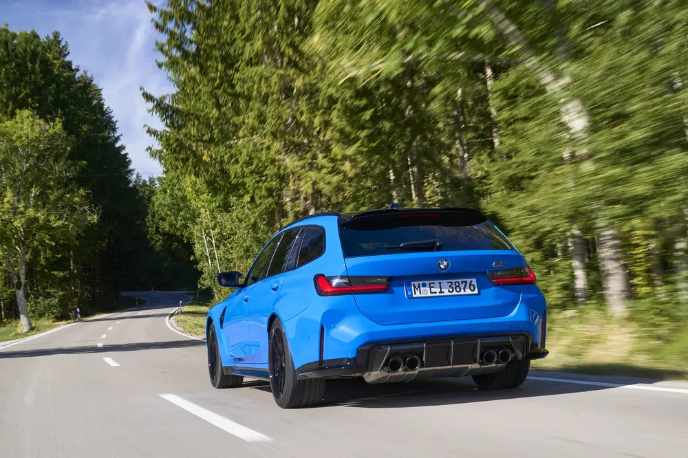 Blauwe BMW M3 Touring rijdend op een boomrijke weg, dynamisch achteraanzicht.