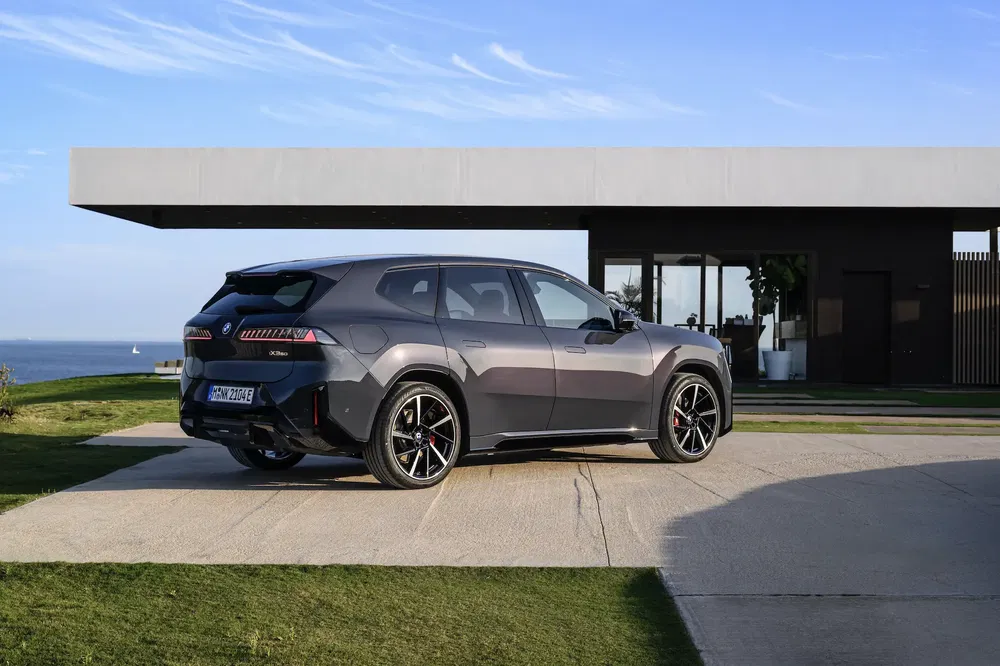 BMW iX électrique garée devant une maison moderne avec vue sur mer, ciel bleu en arrière-plan.