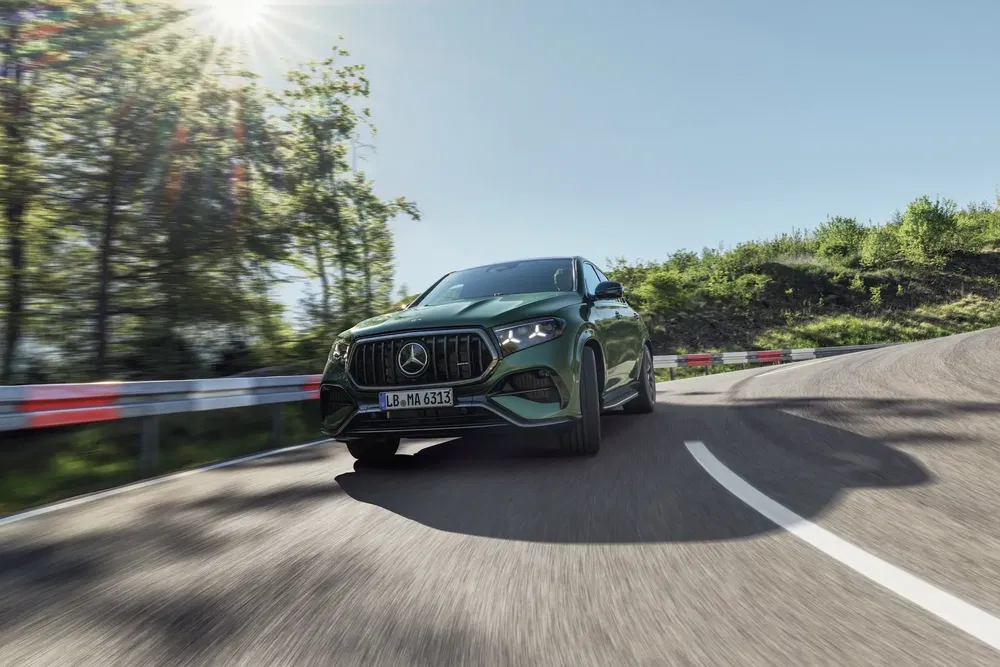 Mercedes-Benz vert sur une route sinueuse ensoleillée entourée d'arbres verts.