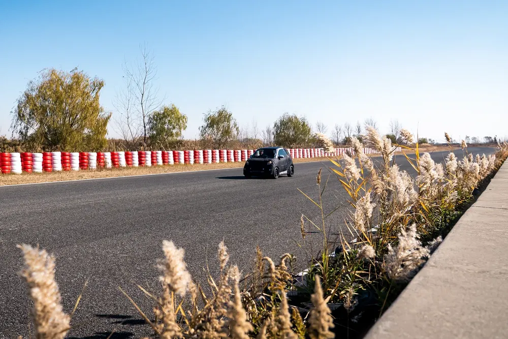 Voiture noire en mouvement sur circuit, bordée de barrières rouges et de végétation sèche.