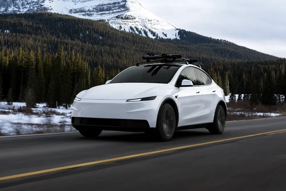 SUV électrique blanc, barres de toit, roulant sur route de montagne enneigée, forêt en arrière-plan.