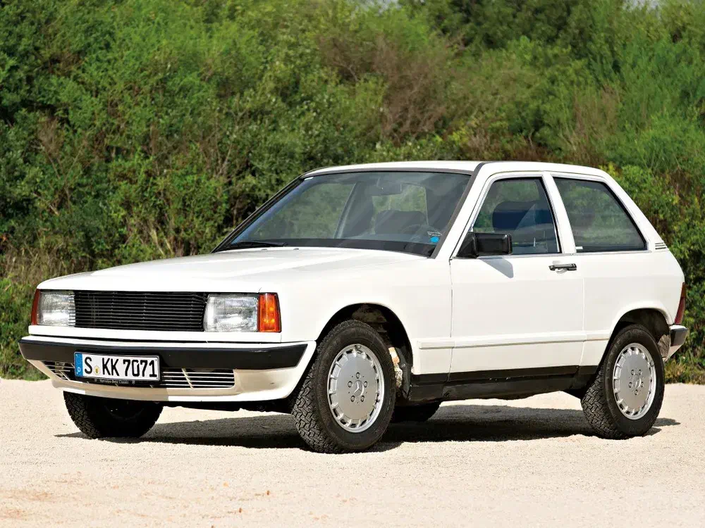 Ford Fiesta 1980, blanche, garée sur un chemin gravillonné, fond d'arbres verts.