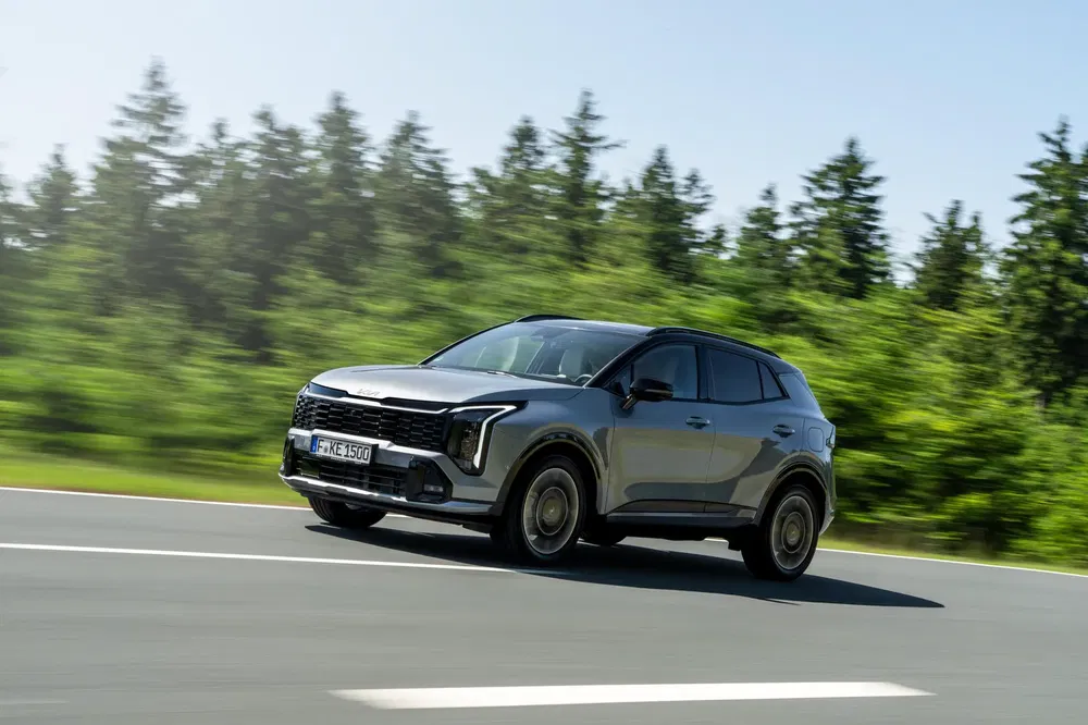 SUV Kia roulant sur une route bordée d'arbres, ciel dégagé, véhicule en mouvement.