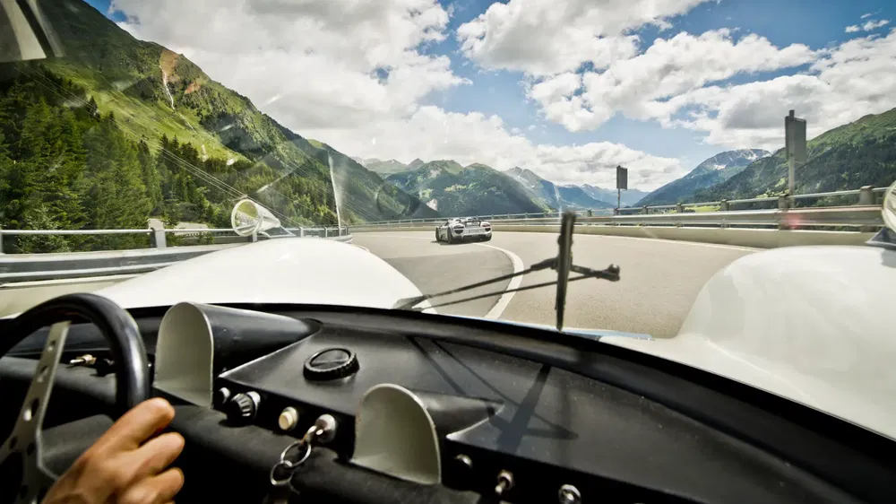Vue intérieure d'une Porsche sur une route panoramique de montagne, ciel bleu et nuages.
