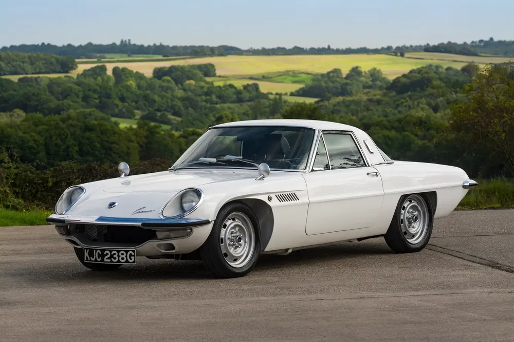 Mazda Cosmo Sport blanc vintage sur une route de campagne, paysage verdoyant.