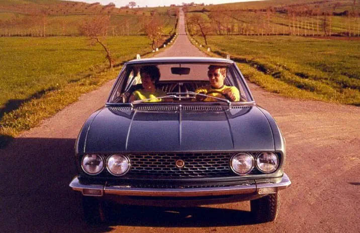 FIAT 124 Sport Coupé parcourant une route pittoresque avec deux passagers en avant-plan.