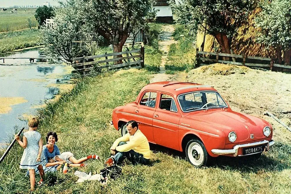 Renault Dauphine rouge garée près d'un étang, famille pique-niqueant en milieu rural.