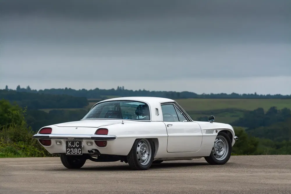 Mazda Cosmo Sport blanche stationnée devant un paysage verdoyant et ciel nuageux.