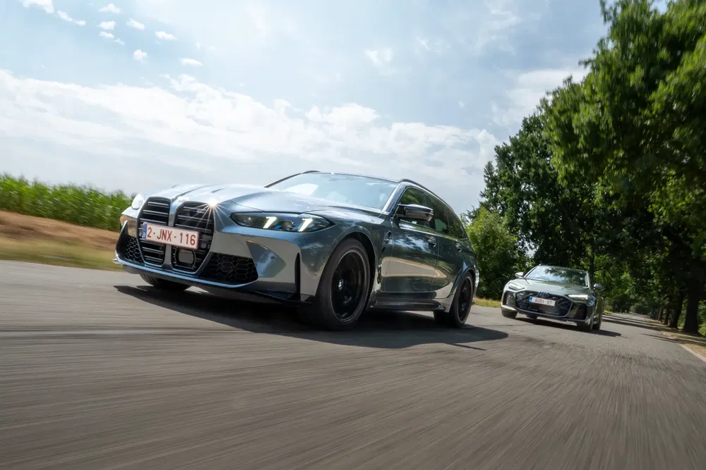 Twee zilveren BMW M3 Touring rijden op een met bomen omzoomde weg.