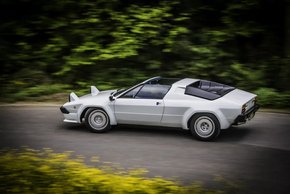 Lamborghini Silhouette blanche, voiture sportive classique, roulant sur une route bordée d'arbres.