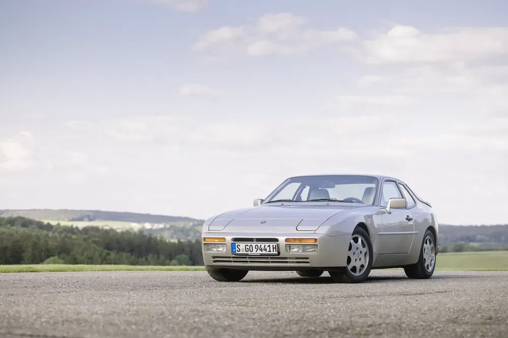 Porsche 944 grise stationnée sur une route, paysage champêtre en arrière-plan.